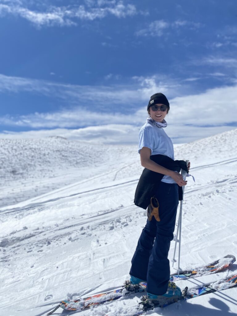 Lexi Milan wears black ski pants and a blue t-shirt with a black ski jacket tied around her waist. She wears a black hat and sunglasses and is standing on skiis on a snowy mountain with a sunny background.