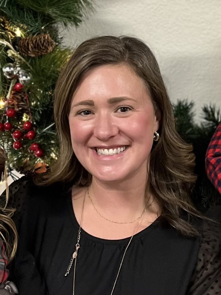 Rachel Jorgensen has long curled brown hair and is wearing a black shirt and a gold necklace. She is standing in front of a holiday garland.